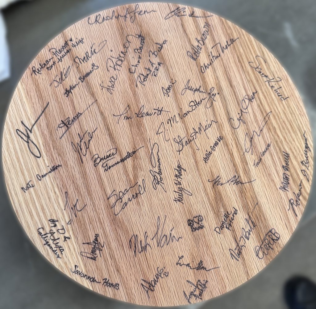 Bottom view of red oak circular cribbage board featuring numerous handwritten signatures and natural wood grain, part of a custom retirement gift.