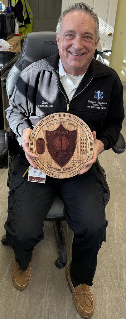 Tom proudly holding his custom circular cribbage board featuring a red oak base, Padauk Stewart’s Ambulance shield inlay, engraved scoring track, and personalized retirement details.