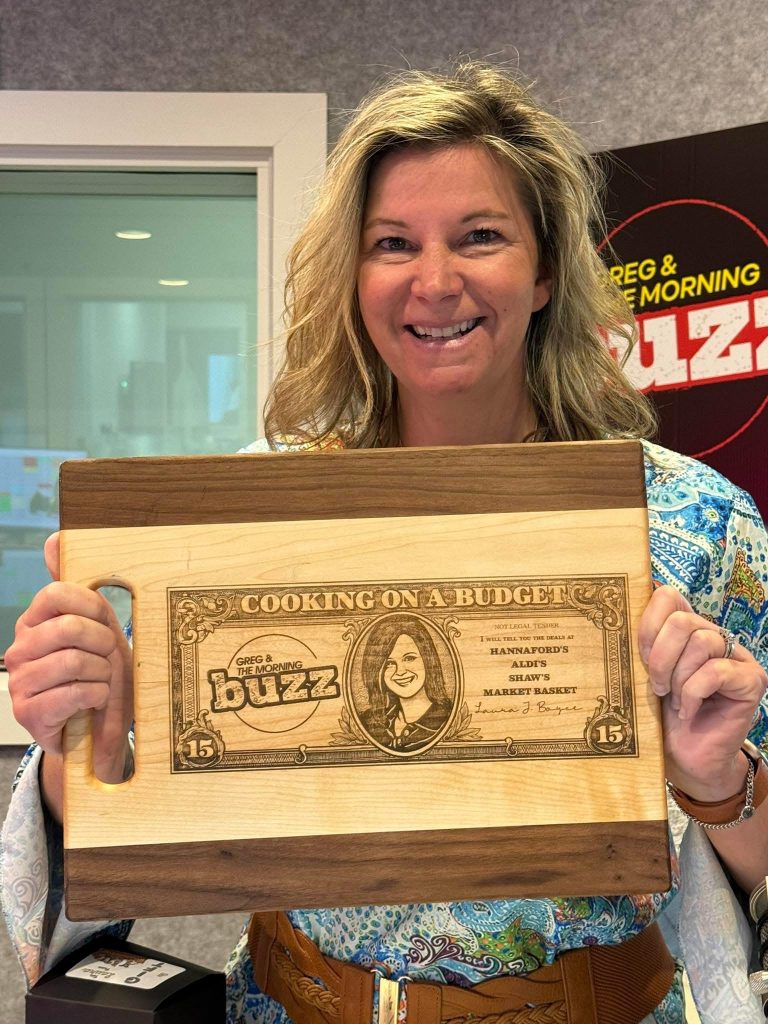 Laura from Greg & The Morning Buzz holding the custom “Cooking on a Budget” walnut and maple cutting board created by 3 Dogs Woodworking.